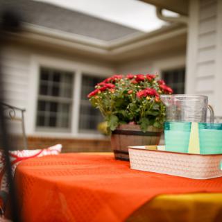 An outdoor table decorated with a vibrant orange tablecloth, featuring a potted plant with red flowers and stacks of books alongside glasses.
