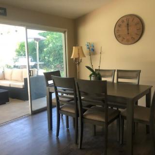 A cozy dining area with a wooden table and chairs, featuring a wall clock and an indoor plant, next to a glass sliding door leading to an outdoor patio.