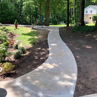 A curving concrete pathway winds through a landscaped garden area surrounded by lush greenery and trees.