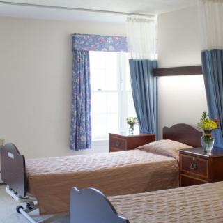 A hospital room with two patient beds, a window with blue curtains, and wooden furniture adorned with flowers.