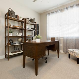 A cozy home office with a wooden desk and a bookshelf, filled with decor items, illuminated by natural light from a large window.