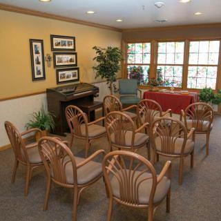 A small meeting room with wooden chairs arranged facing a table and piano, adorned with decorative frames and indoor plants.