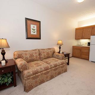 A cozy living room with a patterned sofa, two lamps, and a small kitchenette in the background.