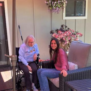A young woman shares a moment with an elderly person in a wheelchair, surrounded by flowers on a sunny day.
