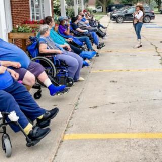 A group of people seated in wheelchairs outdoors, listening to a person speaking in a parking lot.