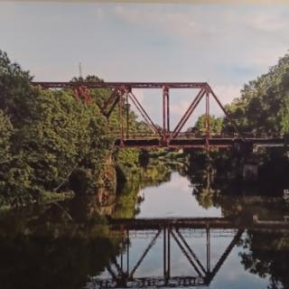 A serene view of a red metal bridge reflecting in the calm waters of the river, surrounded by lush greenery.