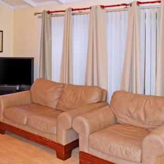 A cozy living room with two beige sofas, a television in the corner, and light-colored curtains covering the windows.