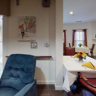 A cozy living room with a comfortable chair and a dining area featuring tables with floral arrangements.
