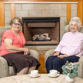 Two elderly women sitting comfortably by the fireplace with cups on the coffee table.
