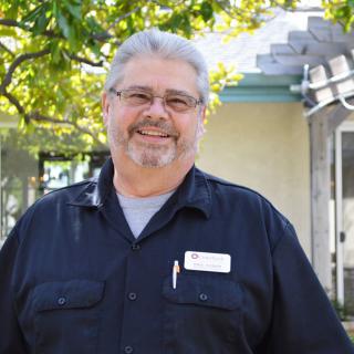 A man smiling outdoors, wearing a name tag and casual clothing.