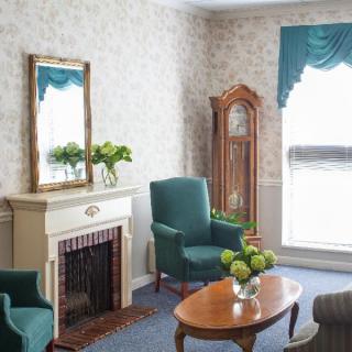 A cozy living room with traditional decor, featuring a fireplace, green armchairs, a grandfather clock, and a wooden coffee table.
