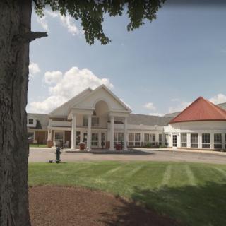 A large building with a grand entrance, surrounded by well-maintained lawns and a tree, set against a backdrop of blue sky with clouds.
