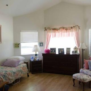 A cozy bedroom with light-colored walls, a bed, dresser, chair, and window decorations.