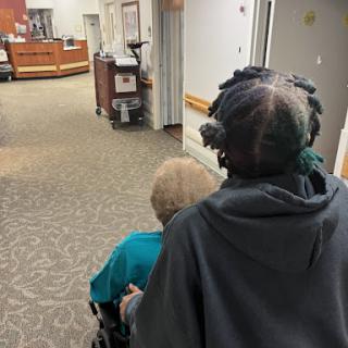 A caregiver pushing an elderly person in a wheelchair through a healthcare facility.