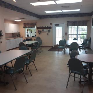 A spacious community center meeting room with tables and chairs, featuring a kitchen area and large windows.
