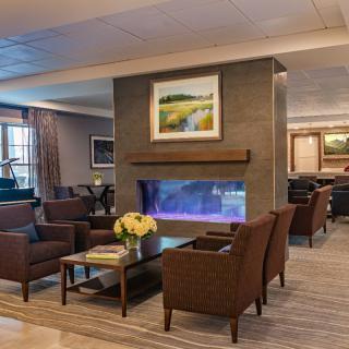A cozy hotel lobby with modern decor, featuring a seating area around a central fireplace and elegant furnishings.