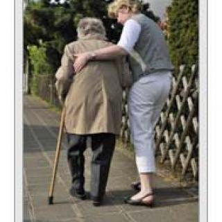 A person assisting an elderly individual with a cane while walking outdoors.