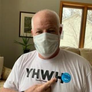 A person wearing a mask and a t-shirt with 'YHWH' printed on it, pointing to a sticker while indoors.
