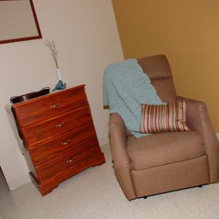 A cozy corner featuring a comfortable chair with a throw blanket and a striped pillow, alongside a wooden drawer chest.