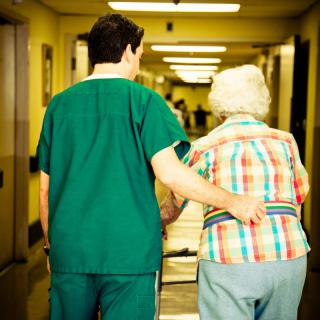 A healthcare worker assists an elderly person down a hospital corridor.