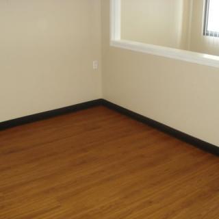 An empty room with wooden flooring and beige walls, featuring a window for natural light.