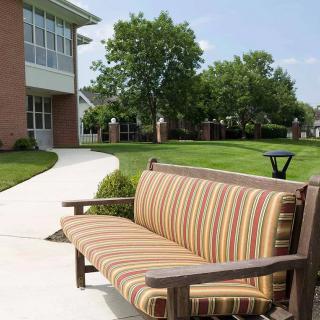 A striped cushioned bench sits along a pathway near a building, surrounded by grass and trees.