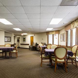 A spacious dining room with tables and chairs arranged neatly, featuring carpeted flooring and large windows allowing natural light.