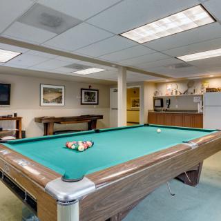 A well-lit recreation room featuring a pool table and a small kitchenette in the background.