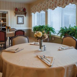 A neatly set dining table in a cozy room with large windows and decorative plants.