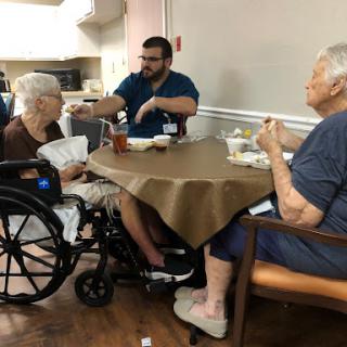 A healthcare worker engages with two elderly residents during meal time in a nursing home.