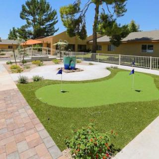 A neatly landscaped outdoor area featuring a mini golf putting green surrounded by trees and benches.
