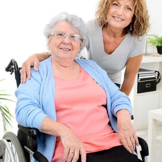 An elderly woman in a wheelchair smiling with her caregiver beside her indoors.