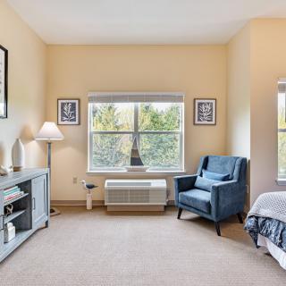 A cozy bedroom with a large window, featuring an armchair, a lamp, and decorative elements.