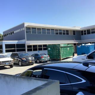 A view of an office building with a parking lot filled with cars and dumpsters during the day.