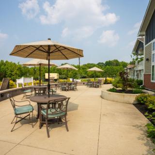 A spacious patio with tables and umbrellas, surrounded by greenery and adjacent to a building.