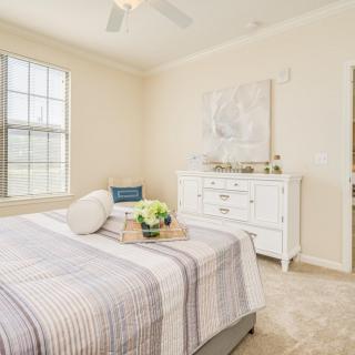 A cozy bedroom with neutral tones featuring a made bed, a dresser, and large windows.