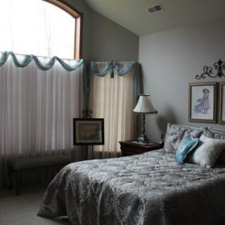 A cozy and elegant bedroom featuring a neatly made bed, soft color palette, and natural light streaming through large windows.
