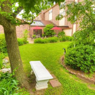 A serene garden scene with a white bench under a tree, surrounded by lush greenery and a view of a patio with red umbrellas.