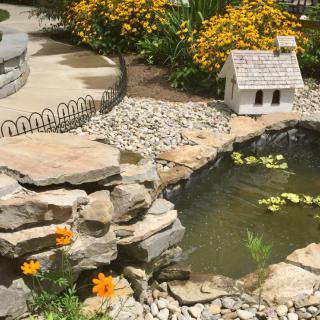 A serene garden setting featuring a pond with water lilies, surrounded by rocks and vibrant flowers, along with a decorative miniature house.