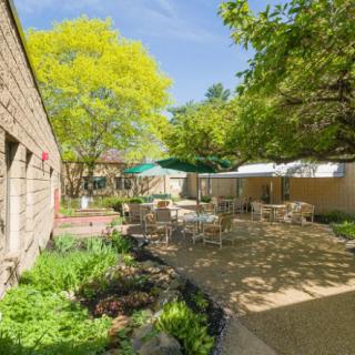 A tranquil courtyard featuring outdoor seating with tables and chairs, surrounded by lush greenery on a sunny day.