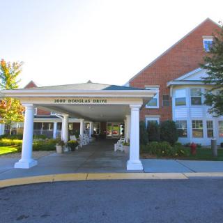 A building entrance with columns and a driveway, surrounded by landscaping and bathed in sunlight.