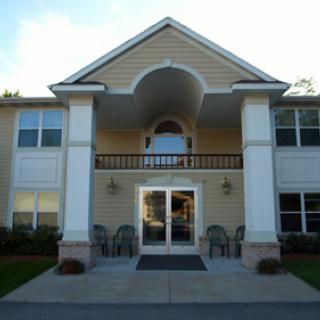 The image shows the front entrance of a two-story residential building with columns and patio chairs.