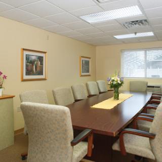A well-lit conference room featuring a long wooden table surrounded by upholstered chairs and a floral centerpiece.