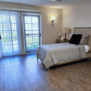A modern bedroom with a neatly made bed, wooden flooring, and large windows allowing natural light.