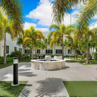 A serene courtyard with a fountain surrounded by palm trees and modern buildings under a bright blue sky.