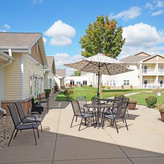 A cozy outdoor patio area with tables, chairs, and an umbrella, surrounded by residential buildings and a tree under a sunny sky.