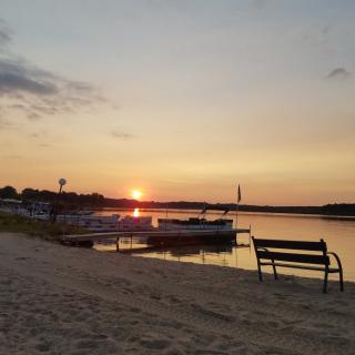 A serene sunset over a lakeside beach with boats and a bench.