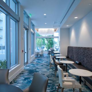 A modern cafe interior with seating areas, featuring multiple tables and chairs, illuminated by natural light through large windows.