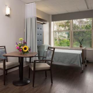 A clean and well-lit hospital room with a bed, table, chairs, and a vase of flowers near a large window.