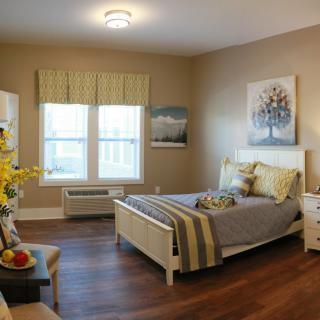 A cozy and well-decorated bedroom with natural light streaming in through two windows, featuring a bed with decorative pillows, wooden flooring, and tasteful wall art.